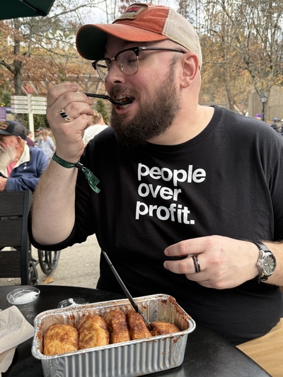 A man with glasses and a beard smiles as he puts something in his mouth.