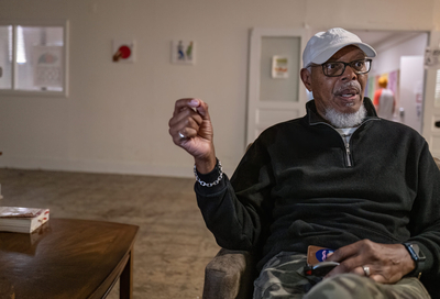 A Black man wearing a white baseball and glasses sits in what appears to be a museum, gesturing with his right hand while he talks.