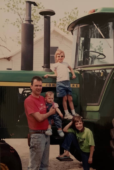 A man stands beside a John Deere tractor holding a baby while two little girls stand on the tractor.