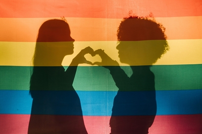 Two women shadowed while making a heart together with their hands behind a LGBTQ rainbow pride flag