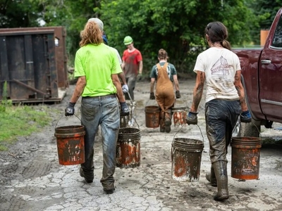 Several people are walking on a muddy path, carrying dirty buckets. They are wearing stained work clothes and boots. Trees and a truck are visible in the background.