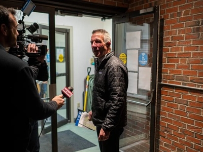 A man stands outside a building speaking to reporters with microphones, holding papers, while a cameraman films.