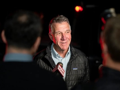 A man in a black jacket speaks to reporters holding microphones outdoors.