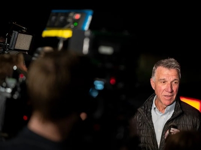 A man in a jacket speaks to media, surrounded by cameras and microphones, in a dimly lit setting.
