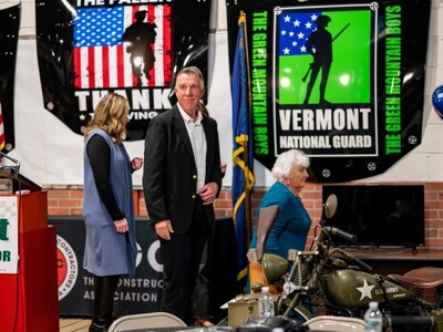 A man and two women stand near a podium in a room with military-themed banners and a vintage motorcycle.