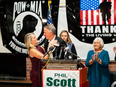 A man is speaking at a podium with several microphones. Banners behind him say "POW-MIA" and "Thank the Living." A woman next to him holds his arm. People in the background are clapping.