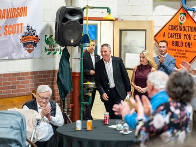 A group of people in a room, some seated and others standing near a Harley-Davidson banner. A man in a suit walks in, greeted by applause. A construction sign is visible on the wall.
