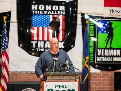 A man speaks at a podium with multiple microphones, standing before banners honoring military service. An American flag is visible to his right.