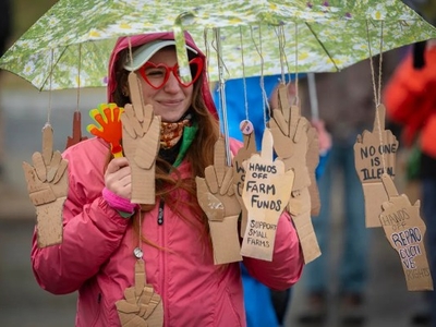 Person with heart-shaped glasses holds an umbrella decorated with cardboard hand cutouts, displaying messages advocating farm funds and reproductive rights.