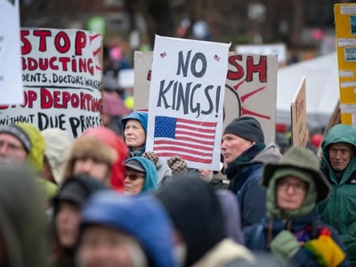 A crowd of protesters holding various signs, including one that reads "NO KINGS!" with an American flag design. People are dressed warmly, suggesting cool weather.