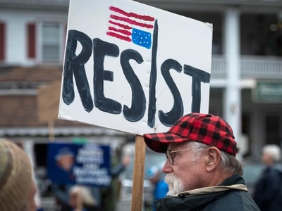 An older man holds a sign reading "RESIST" with an upside-down flag at a protest, standing outdoors near buildings, surrounded by other people.