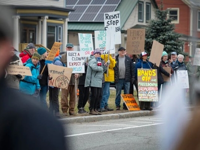 A group of people hold various protest signs on a sidewalk, including messages like "Hands off," "Resist," and "Elect a clown in, expect a circus.
