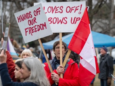 Protesters holding signs that say "Hands off our freedom" and "Gloves off! Elbows up" with a Canadian flag. Crowded outdoor setting with tents and trees in the background.