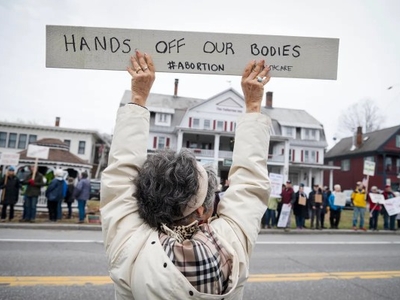Person holds a sign reading "HANDS OFF OUR BODIES #ABORTION" at a protest. A crowd of individuals with signs stands in the background.