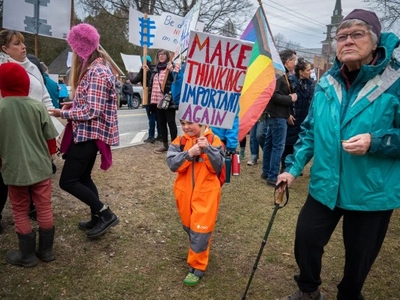 A child holds a sign reading "Make Thinking Important Again" at a protest, surrounded by people carrying various signs.