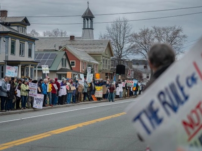 A crowd of people with protest signs line a street, some houses and trees in the background. One sign in the foreground reads, "The Rule of Law NOT the Rule of Man.