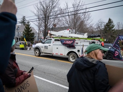 People stand by the roadside with signs. A utility truck with a political banner passes by. Bare trees and residential houses are visible in the background.