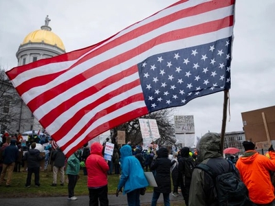 Crowd gathered in a protest, with an American flag flown upside down and people holding signs. The sky is overcast, and a building with a gold dome is in the background.