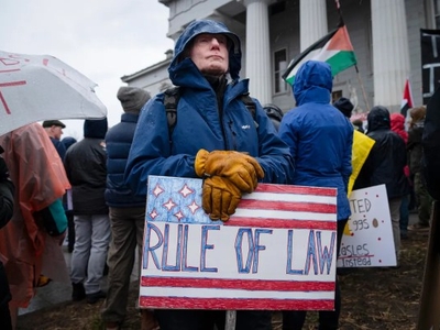 Person in a raincoat and gloves holds a "Rule of Law" sign in a crowd outside a building.