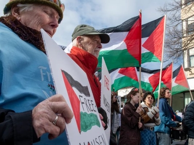A group of people hold signs and Palestinian flags at an outdoor protest, with some signs reading "Free Palestine.