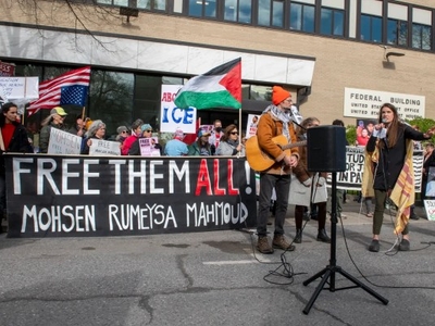 A group of protesters stand outside a federal building holding signs, including a large banner reading "FREE THEM ALL! MOHSEN RUMEYSA MAHMOUD.