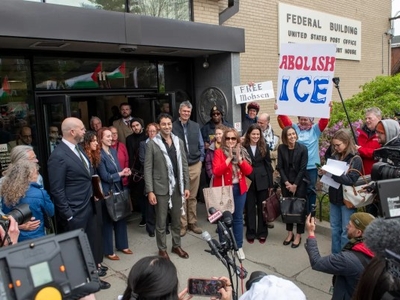 A group of people stand outside a federal building surrounded by media; one person holds a sign reading "ABOLISH ICE," and another displays a "FREE Mohsen" sign.