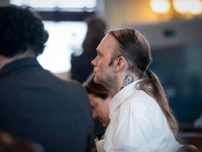 A man with a tattoo on his neck and long hair tied back sits in a courtroom, wearing a white shirt. Other people are seated nearby.
