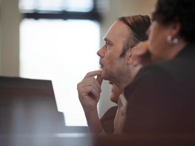 A man sits indoors at a desk, resting his chin on his hand and looking ahead thoughtfully, with another person blurred in the foreground.