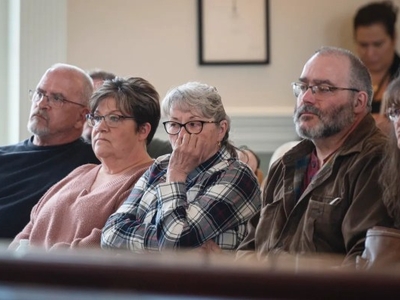 Five adults sit in a courtroom, appearing serious and attentive; one woman covers her mouth with her hand.