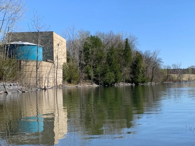 A beige building with a blue section stands by the edge of a calm lake, surrounded by trees under a clear blue sky.