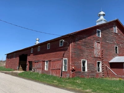 A large, weathered red barn with two cupolas stands next to a rural road under a clear blue sky.