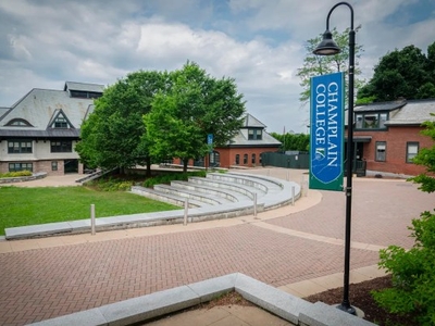 Campus courtyard with brick paths and buildings, a row of trees, and a lamppost displaying a blue "Champlain College" banner.