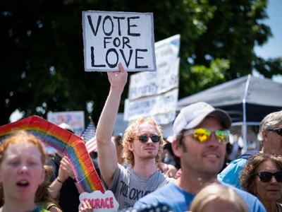 A crowd of people at a rally; one person in the center holds a sign that reads “VOTE FOR LOVE.” Other signs and a rainbow decoration are visible.