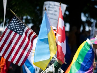 Several people hold flags including the United States, Ukraine, Canada, and a rainbow pride flag at an outdoor gathering.
