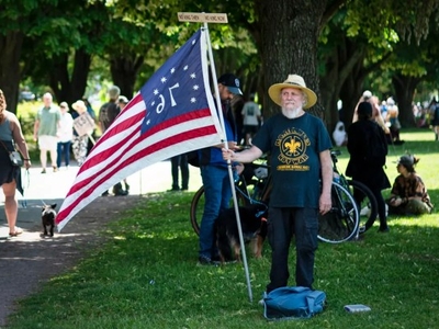 An older man stands in a park holding an American flag with symbols on it. People and trees are in the background.