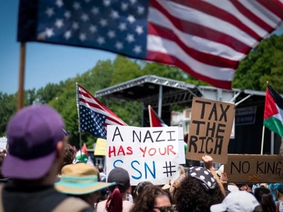 A crowd at a protest holds signs reading “A Nazi has your SSN #” and “Tax the rich” with American and Palestinian flags visible.