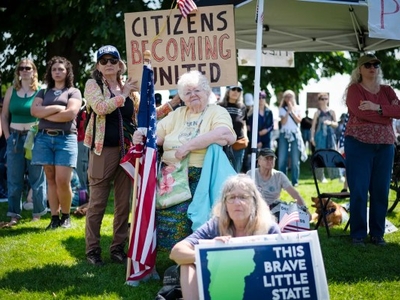 A group of people participate in a rally. One woman holds a sign reading "Citizens Becoming United" and another displays a sign that says "This Brave Little State.
