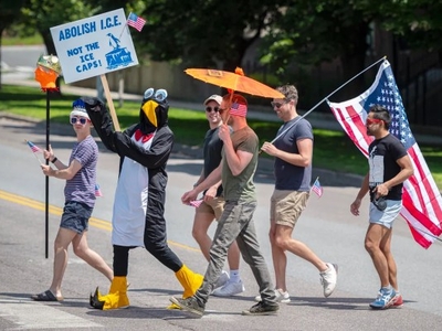 Five people walk on a street; one in a penguin costume holds a sign reading “Abolish I.C.E.—Not the Ice Caps!” Others carry American flags and an orange umbrella.