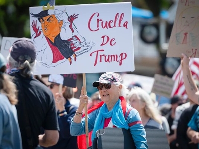 A woman at a protest holds a sign with a caricature of Donald Trump labeled "Cruella De Trump." Other people with signs are visible in the background.