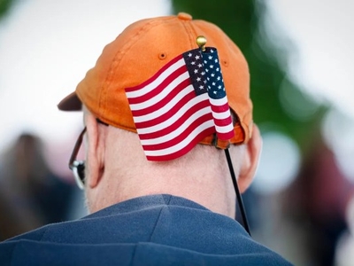 A person wearing an orange cap with a small American flag attached to the back, viewed from behind, with a blurred outdoor background.