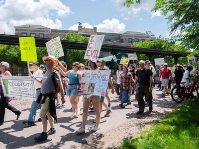 A group of people march outdoors holding various protest signs on a sunny day, with trees and a building visible in the background.