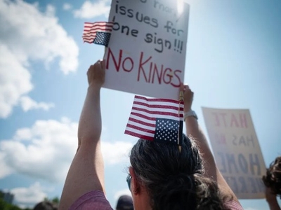 A person holds a sign reading "No Kings" and two small American flags at a protest or rally, with a bright sky in the background.