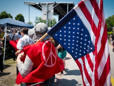 A person wearing a red garment with a peace symbol holds an American flag at an outdoor gathering with people and a stage in the background.
