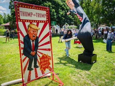 A person swings a bat at an inflatable Donald Trump figure at an outdoor event; a sign reads “Thump-a-Trump” with a cartoon image of Trump and the phrase “Hit him where it hurts!”.