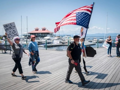 A group of people walk along a marina boardwalk; one person holds a large U.S. flag, another holds a sign reading “This protest is a grief ritual.”.