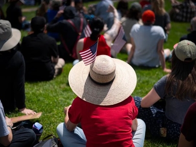 A group of people sit on grass outdoors, many wearing hats and holding small American flags, facing away from the camera.