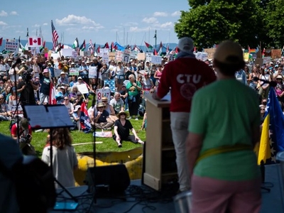 A large crowd gathers outdoors holding signs and flags while listening to speakers on a stage during a public event or protest.