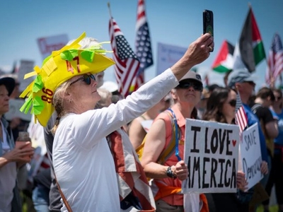 A woman in a yellow hat takes a selfie at a crowded outdoor rally; people around her hold signs and American flags.