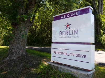 A large sign reads "Welcome Berlin Rehab and Healthcare, 98 Hospitality Drive," standing by a tree-lined road on a sunny day.