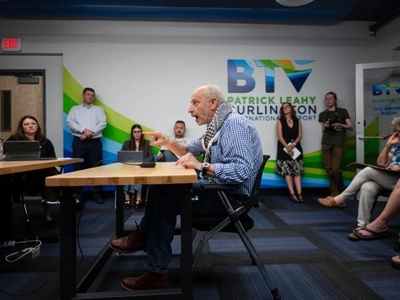 An older man speaks at a table in a meeting room with several people seated and listening. A "Patrick Leahy Burlington International Airport" sign is visible on the wall behind him.